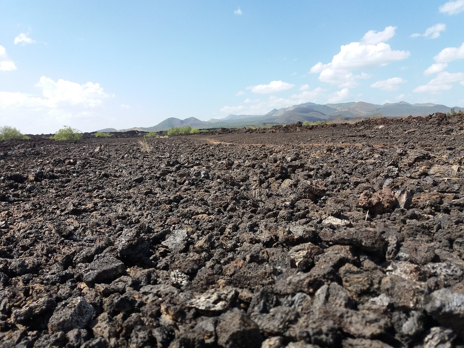 Lavaschotter in Bodennähe fotografiert, im Hintergrund eine Hügelkette unter blauem Himmel