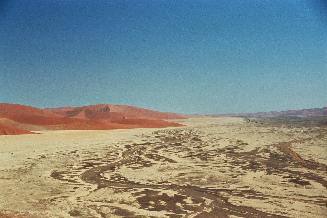 Weite helle Ebene mit dunklen, verzweigten Rinnen im Ton, links rote Dünen, darüber tiefblauer Himmel.