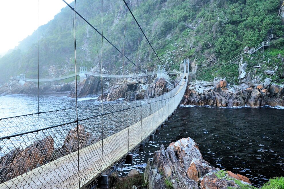 Blick über die Planken der großen Hängebrücke, links dunkles Wasser der Mündung, dahinter zwei kleinere Brücken vor bewaldeten Felshängen
