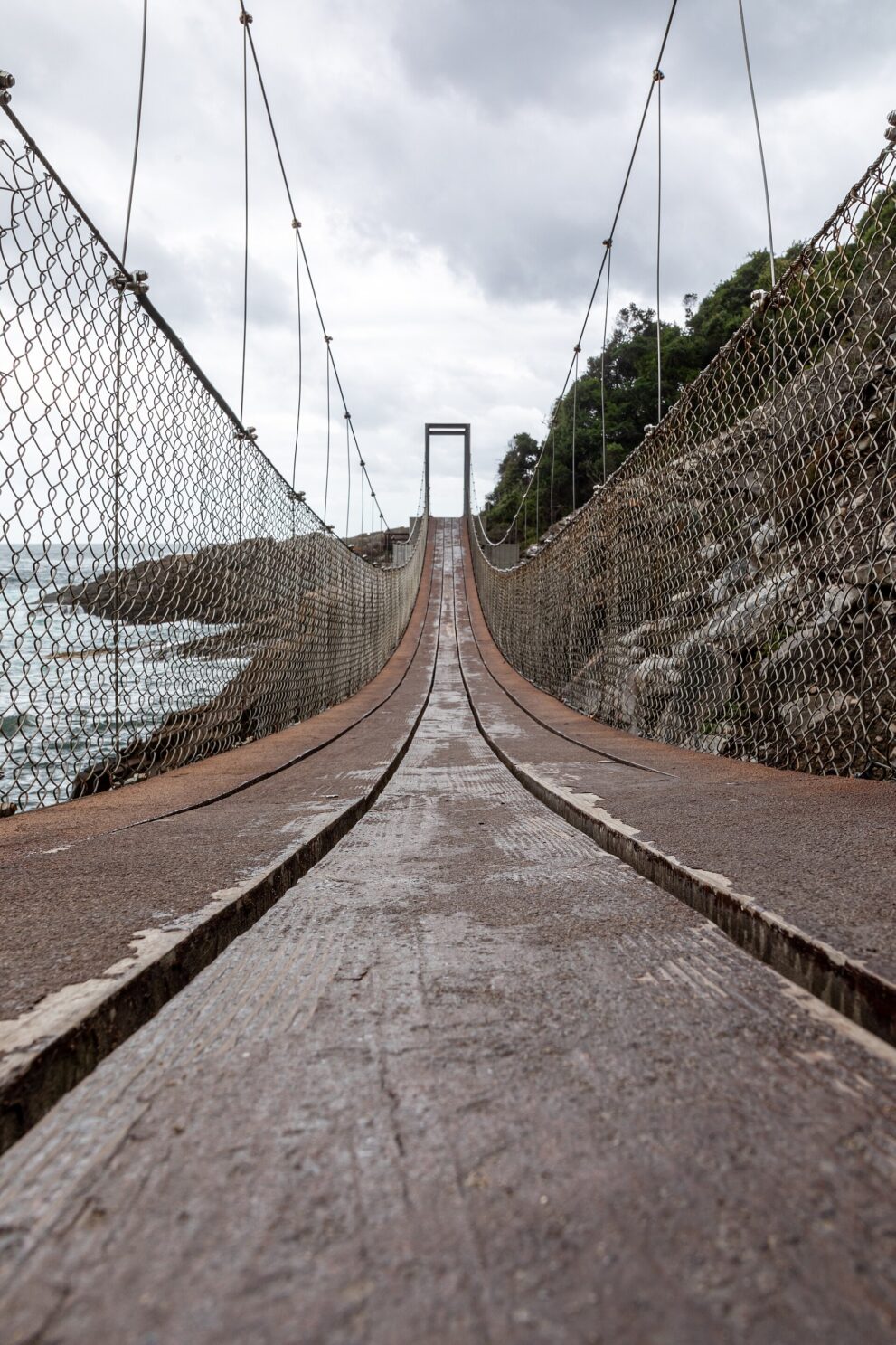 Blick entlang der schmalen Holzplanken einer Hängebrücke zwischen Maschendraht, links Meer und Felsen, am Ende ein Stahlportal