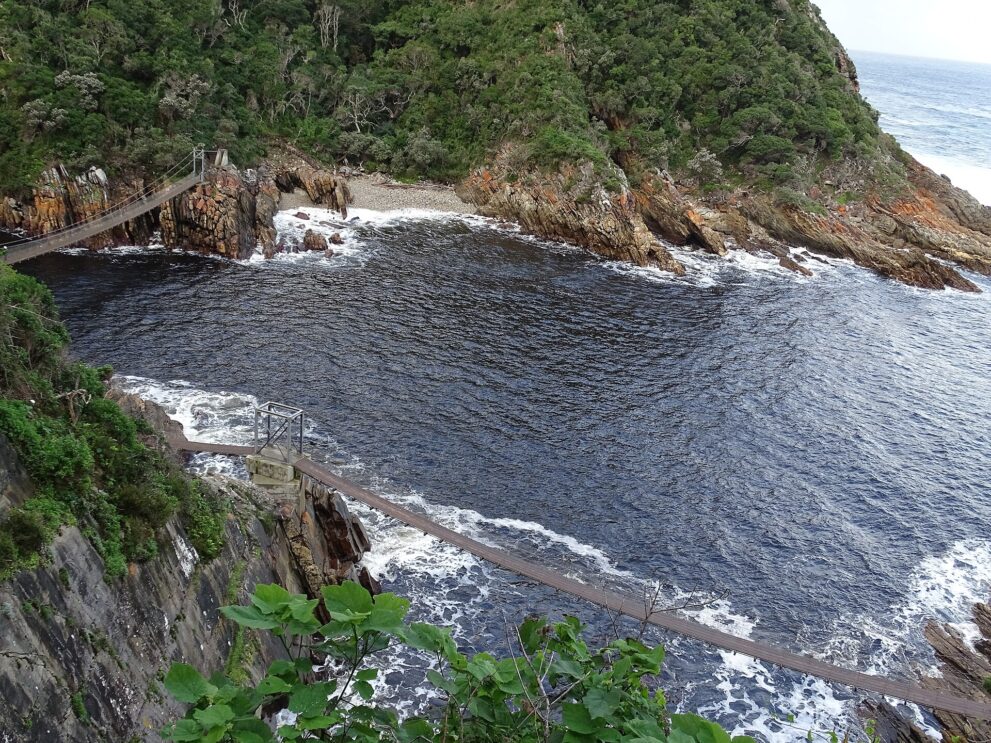 Von oben gesehen: dunkles Wasser der Mündung zwischen gefalteten Felsen, zwei Hängebrücken, im Hintergrund Wald und offenes Meer