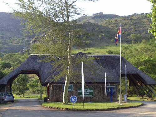 Torhaus mit Strohdach über der Zufahrt, davor Fahnenmast mit der Flagge Eswatinis und grüne Schilder, dahinter ein Berghang