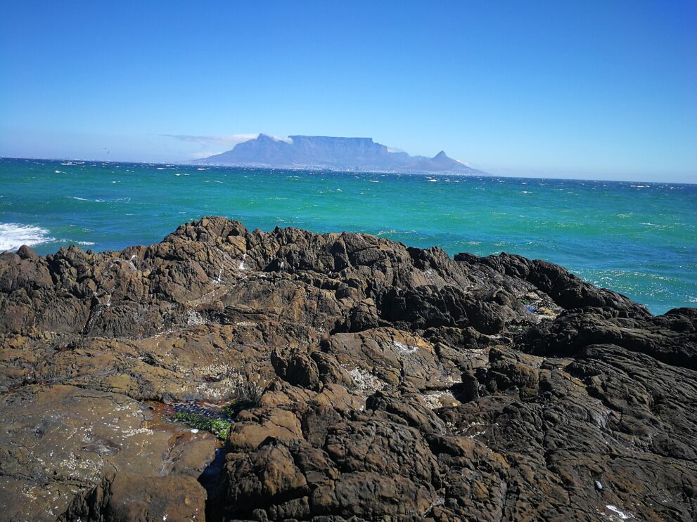 Der flache Tafelberg mit Lion's Head rechts, gesehen über das türkisgrüne Meer, vorn dunkle Uferfelsen