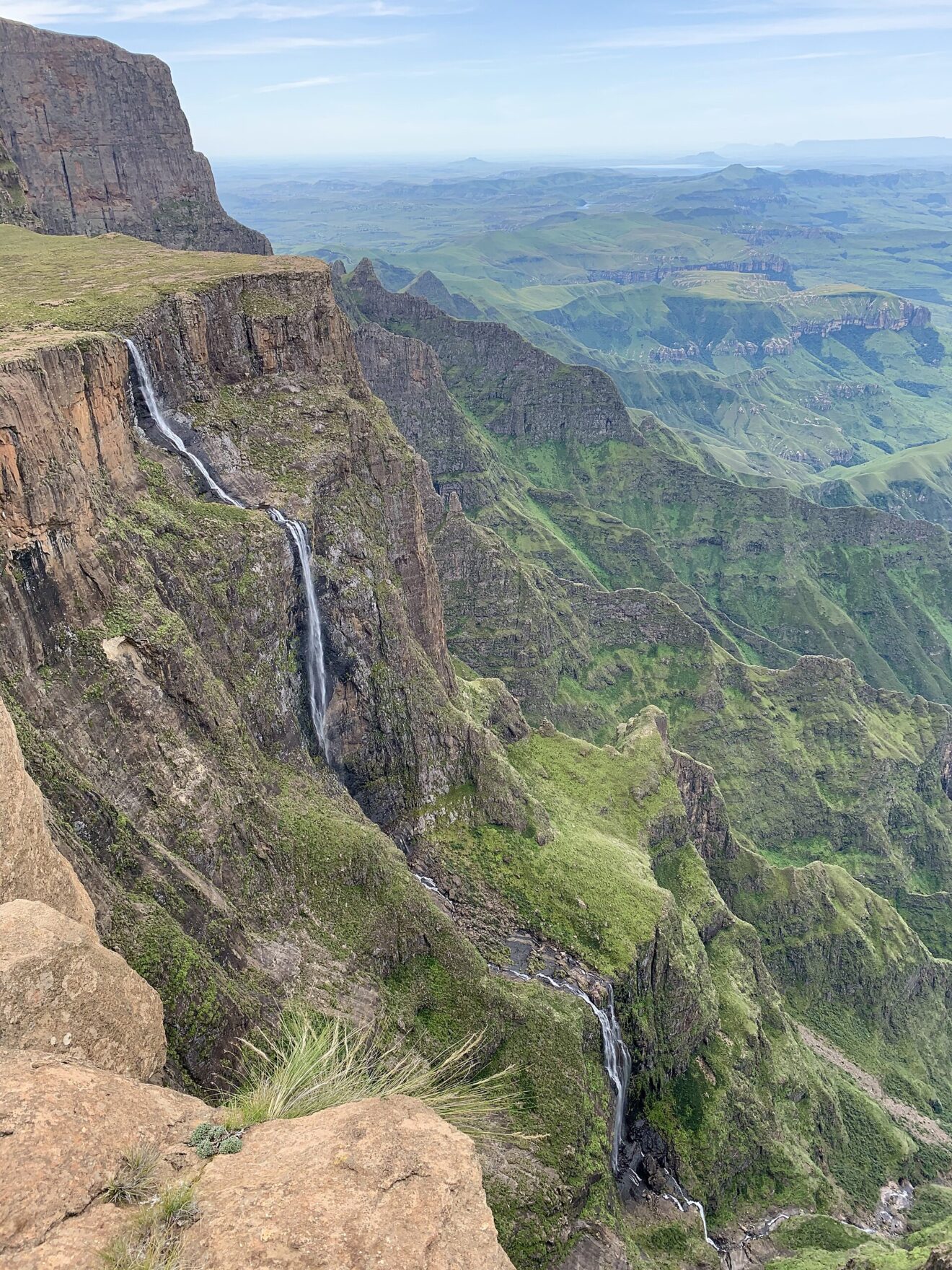 Blick von der Abbruchkante auf den mehrstufigen Wasserfall, der an einer dunklen Felswand in ein grünes Tal fällt.