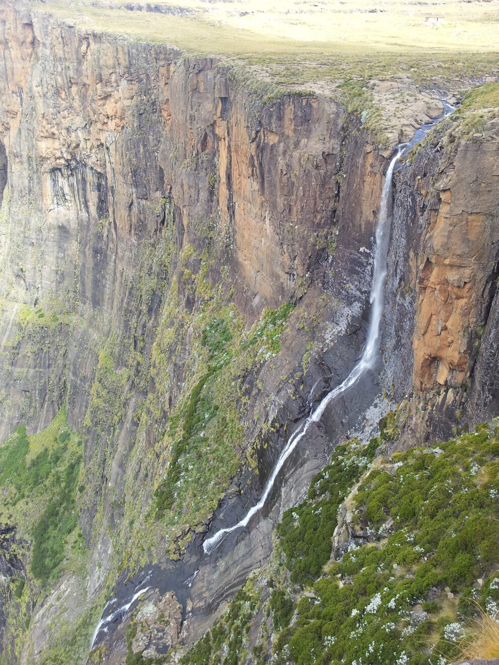 Der Fluss fließt über das grasige Plateau und stürzt als schmales Band über eine senkrechte, rötliche Felswand.