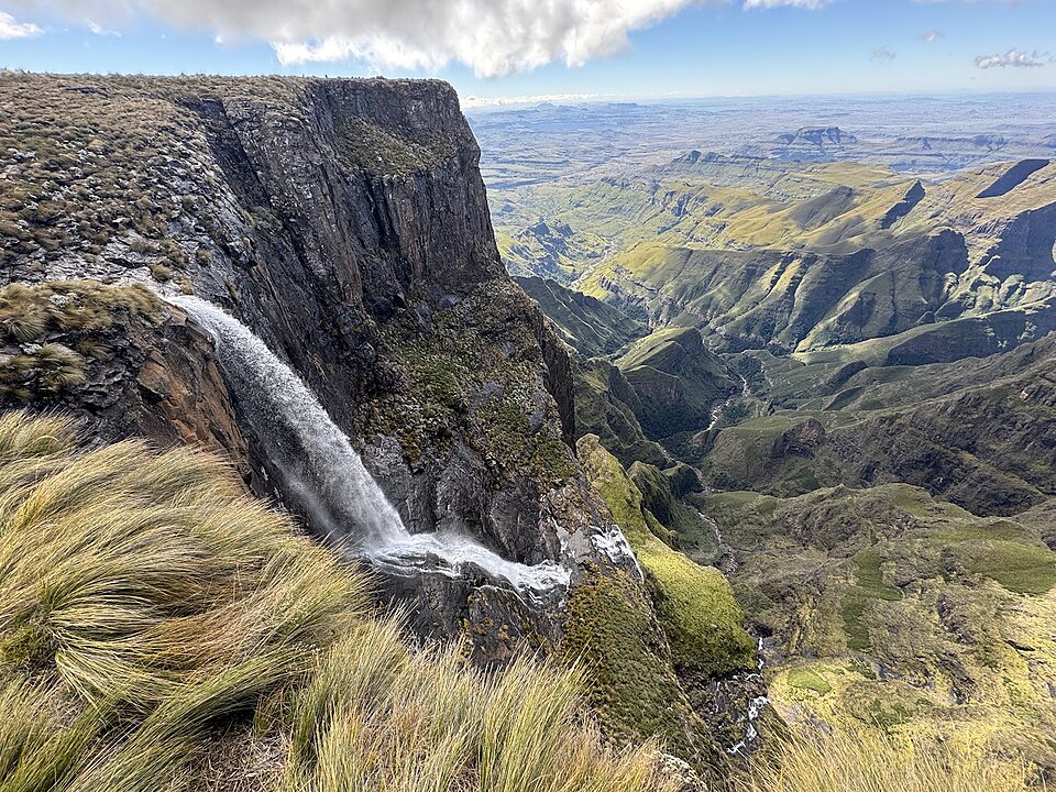 Kräftiger Wasserfall stürzt über eine Felskante in die Tiefe, vorn Grasbüschel im Wind, rechts ein weites grünes Tal.