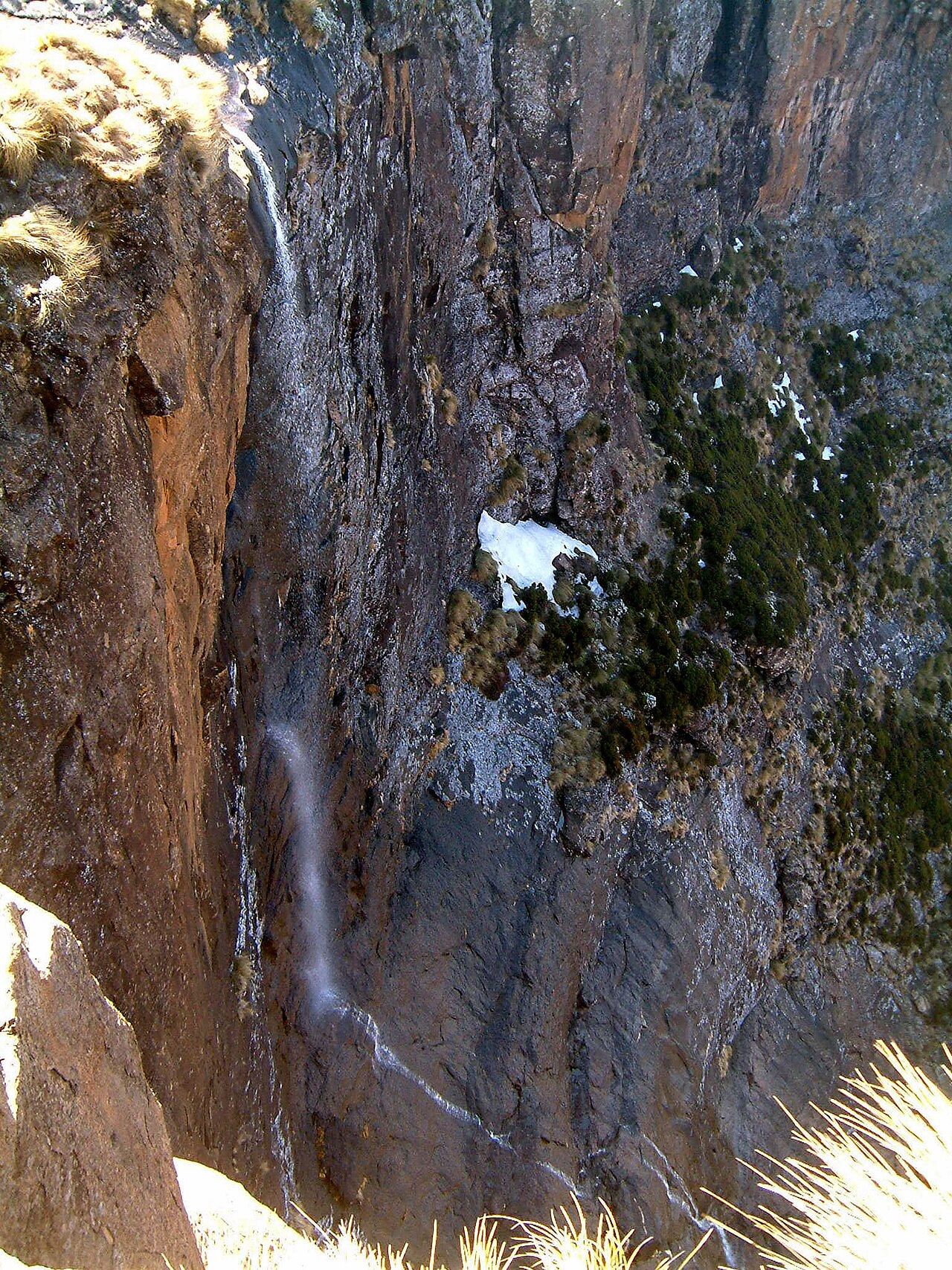 Blick von oben in die Felswand: ein dünner Wasserfaden, daneben Schneereste in der Wand.