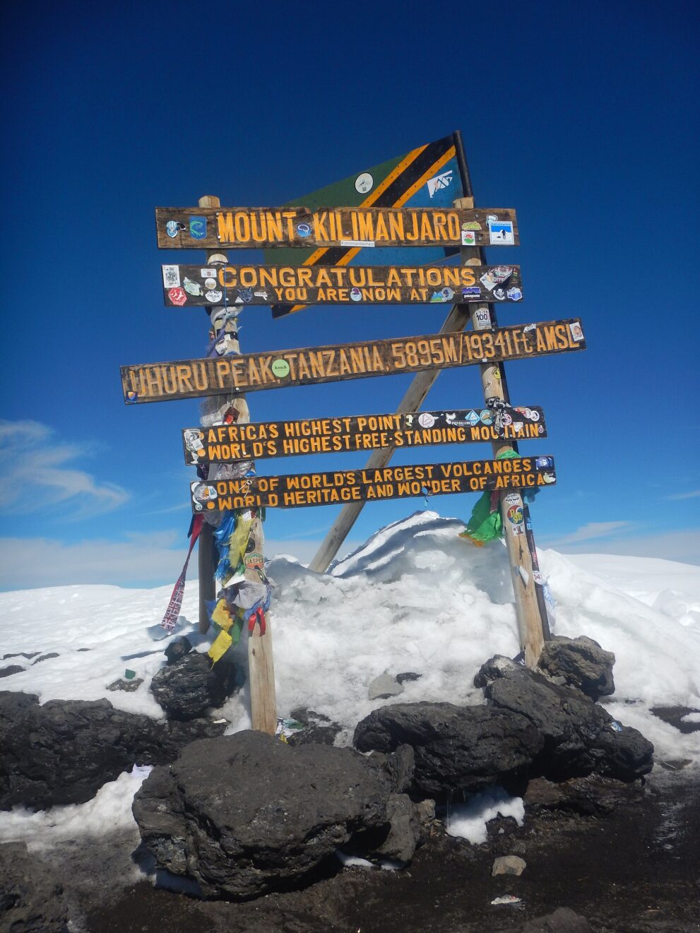 Hölzernes Gipfelschild mit der Aufschrift Uhuru Peak, Tanzania, 5895 m, dahinter Schnee und blauer Himmel