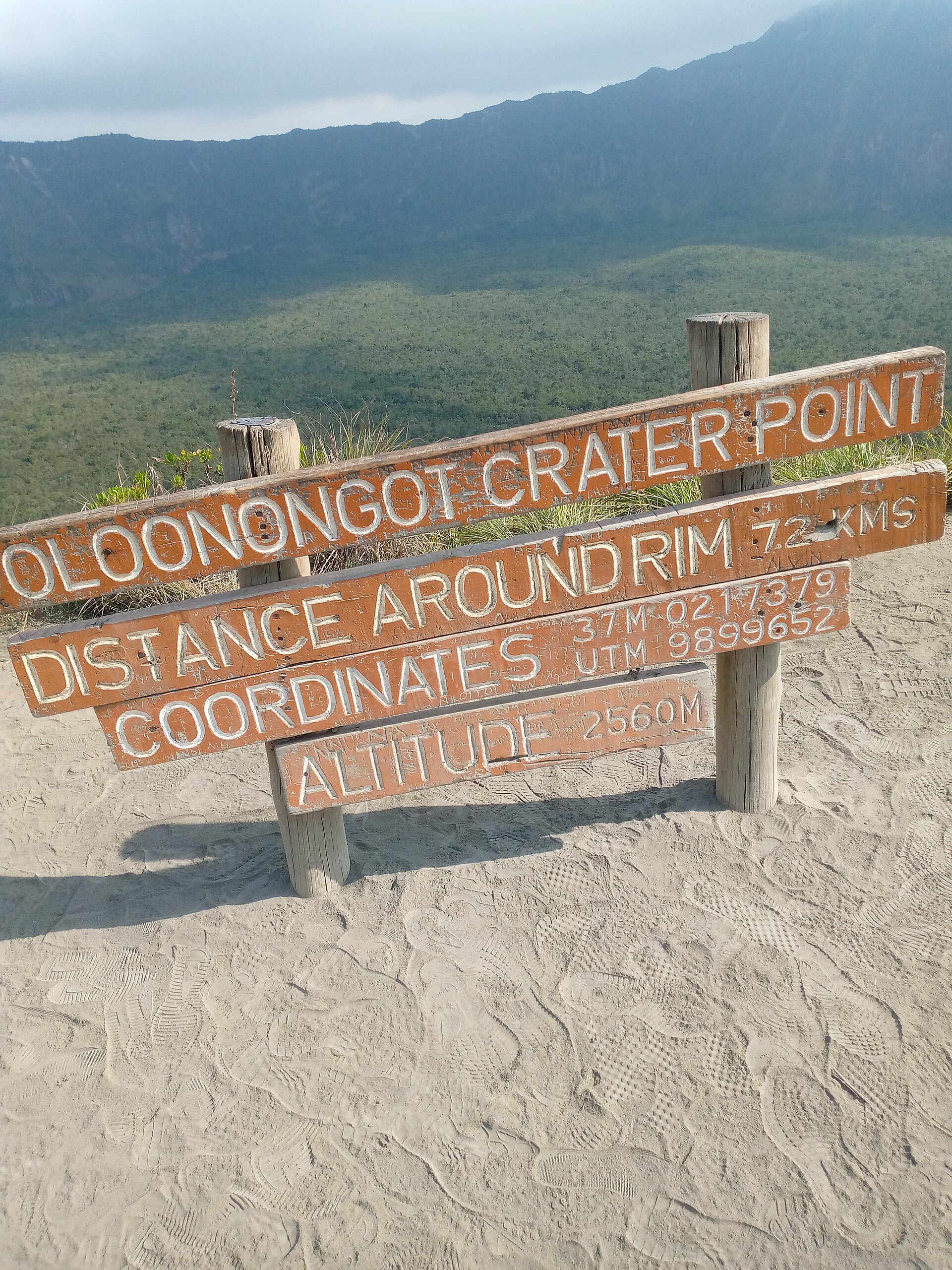Holzschild auf hellem Vulkanstaub voller Fußspuren: Oloonongot Crater Point, Distance around rim 7.2 kms, Altitude 2560 m, dahinter der Krater