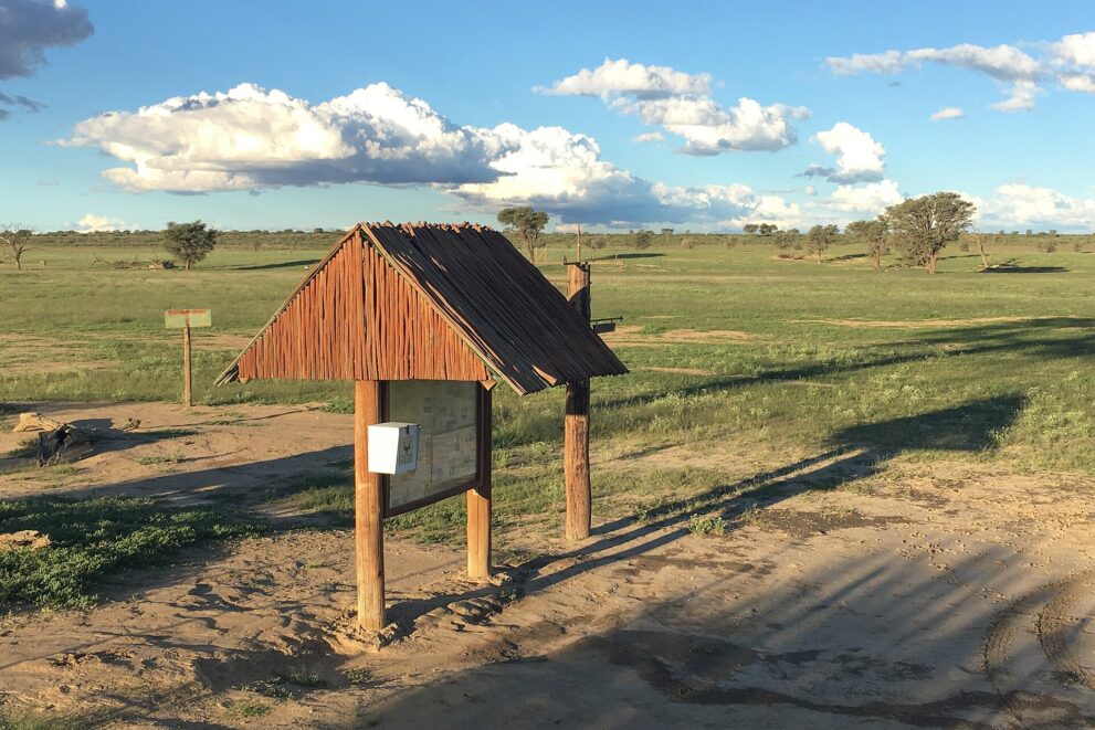 Eine Infotafel unter einem kleinen Reetdach auf sandigem Boden, dahinter eine weite grüne Grasfläche mit einzelnen Akazien