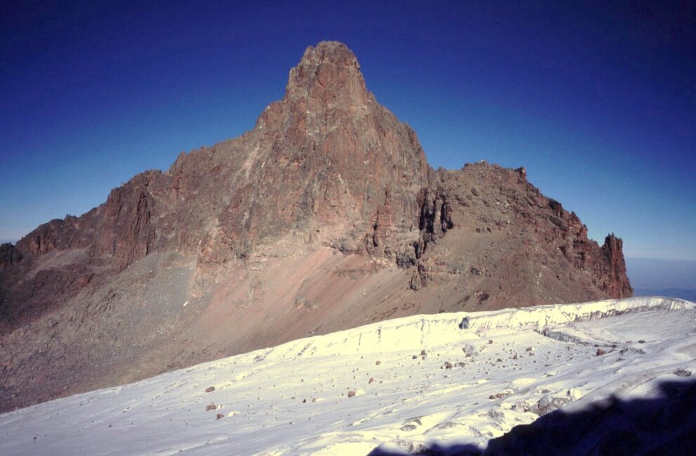 Breite Eisfläche mit verstreuten Steinen und einer Abbruchkante, dahinter der rötliche Felsturm des Nelion vor tiefblauem Himmel