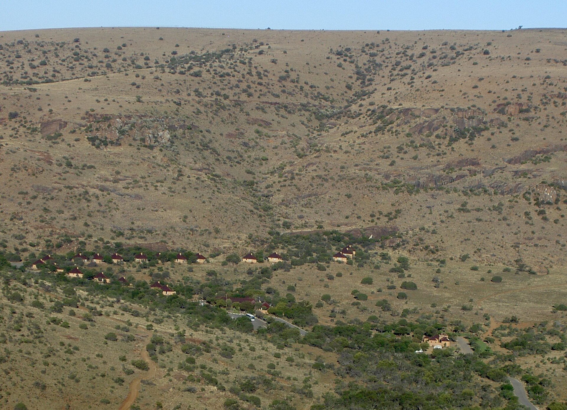 Reihe kleiner Hütten mit roten Dächern im Busch am Fuß eines kahlen, steinigen Hangs, aus erhöhter Lage fotografiert